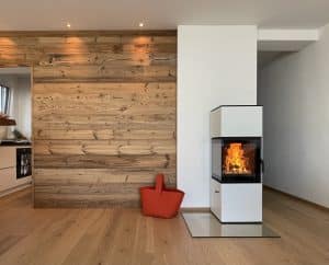 Schlicht eleganter weißer Kamin mit Ecksichtfenster von Ofenmanufaktur auf eckiger Glasplatte in einem Wohnzimmer mit Holz-Rückwand.