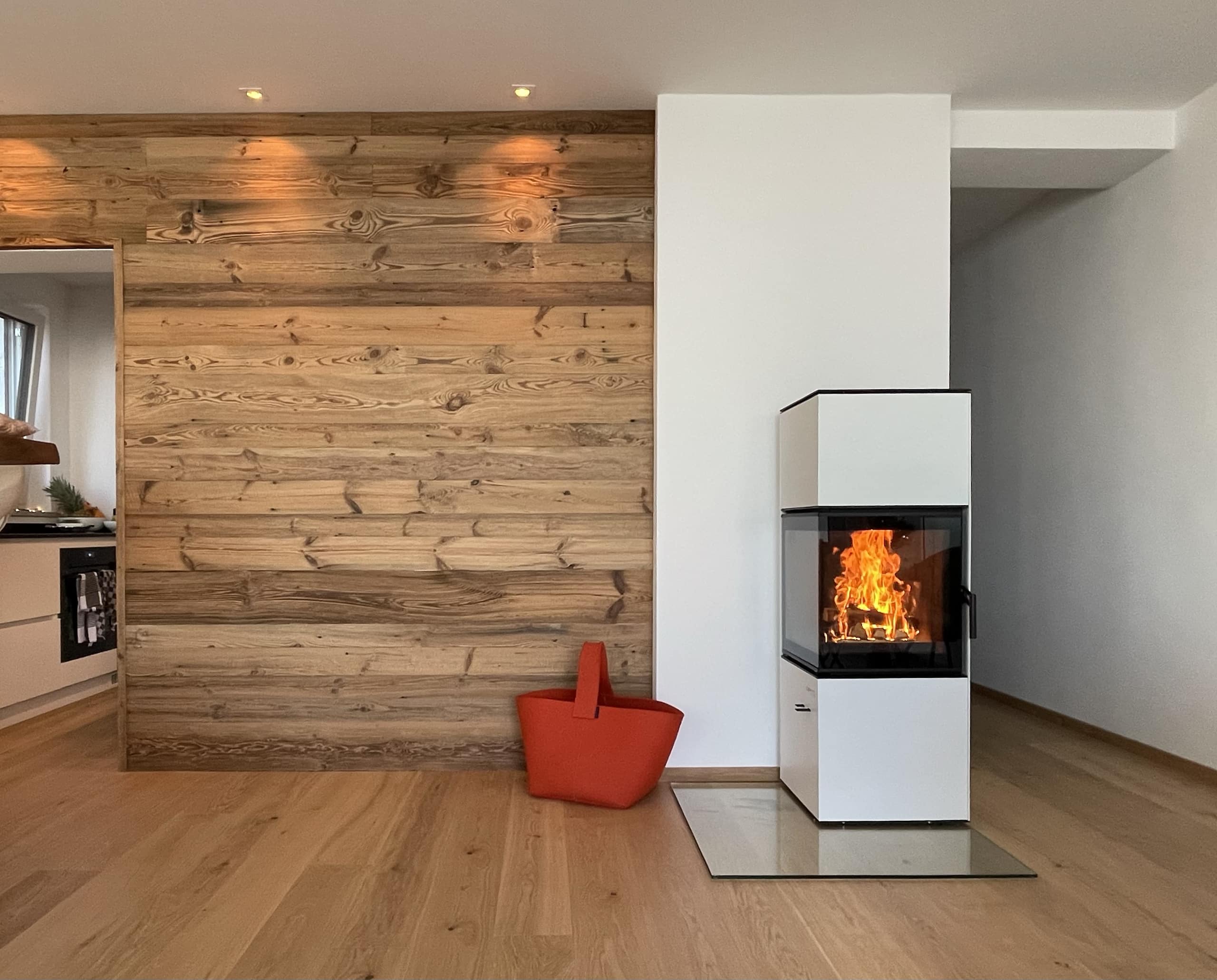 Schlicht eleganter weißer Kamin mit Ecksichtfenster von Ofenmanufaktur auf eckiger Glasplatte in einem Wohnzimmer mit Holz-Rückwand.