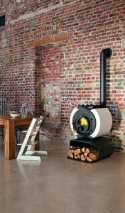 Futuristischer runder Kachelofen aus Gusseisen mit weißen Kacheln auf einem Sockel mit Holzlager von Ofenmanufaktur vor einer Ziegelwand.