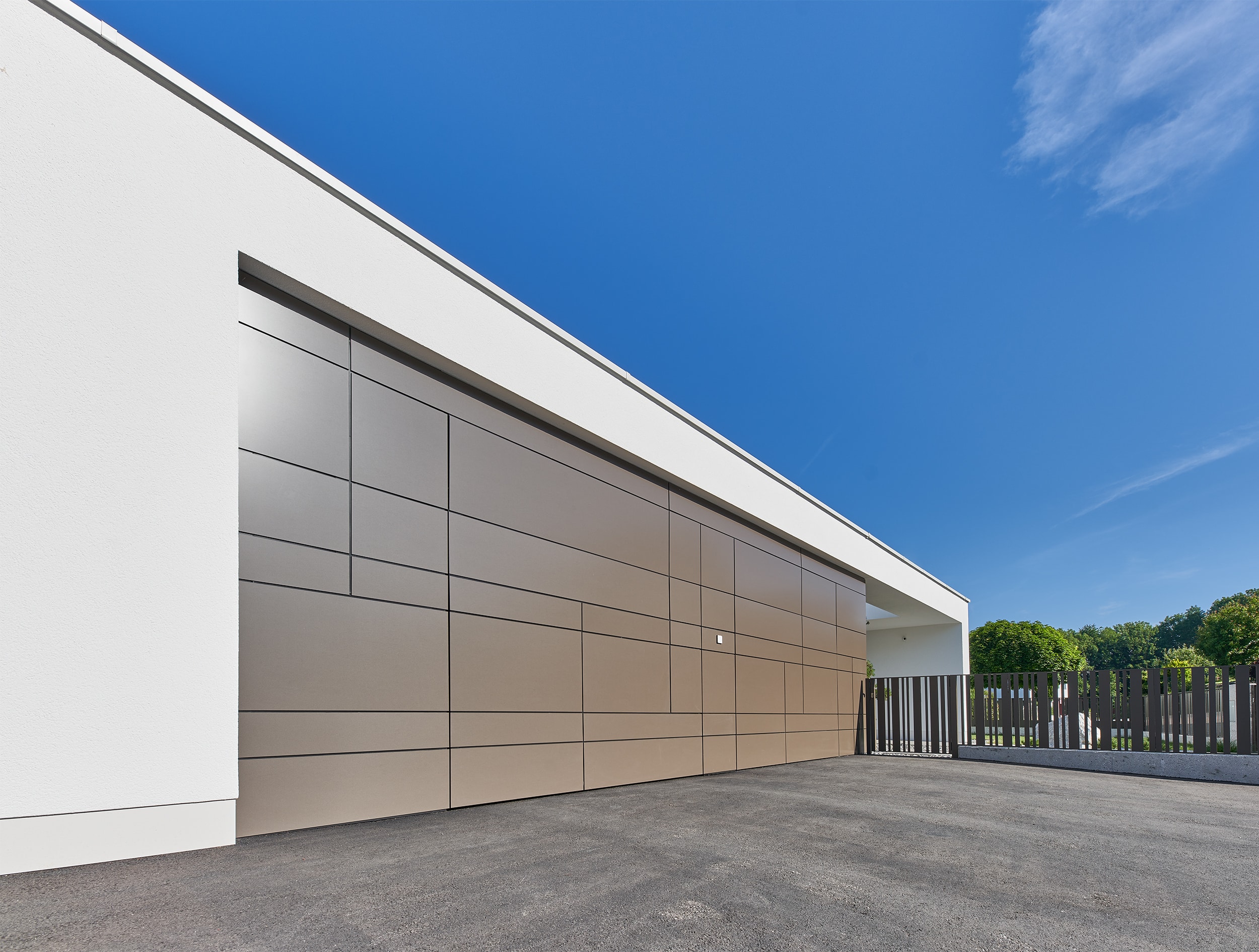 LINDPOINTNER Torsysteme zeigt einen Garagenzubau mit Flachdach und einer asphaltierten Zufahrt zu einem Alu-Rahmen-Tor in braun.