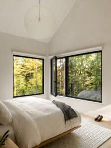 Modernes Schlafzimmer mit großflächiger Fixverglasung und Fenster mit schwarzem Rahmen und herrlichem Ausblick in die Wälder von M SORA.