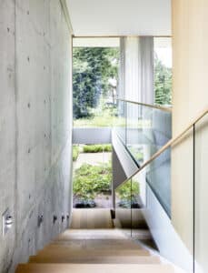 Moderne Holztreppe mit Sichtbetonwand, Glasgeländer und großzügiger Fixverglasung im Galeriebereich mit Gartenausblick von M SORA.