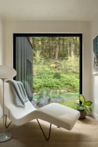 Modernes Wohnzimmer mit großer Fensterfront als Fixverglasung mit schwarzem Rahmen gemütlicher Liege und Blick ins Grüne von M SORA.