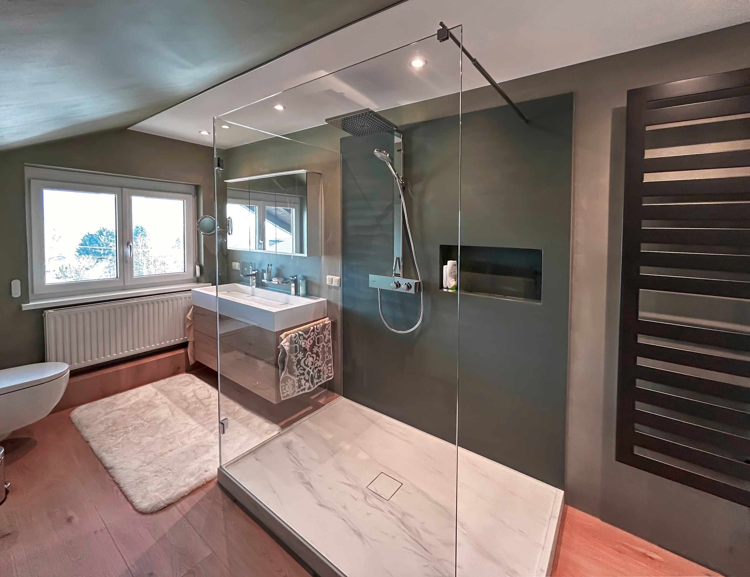 MANO Bad verbindet hier Beton und Mineralwerkstoffe in einer zweiseitigen Glasdusche mit einem schwebendem Waschtisch aus Echtholz und einem schwarzen Handtuchwärmer zu modernem, zeitlosen Badezimmerdesign.