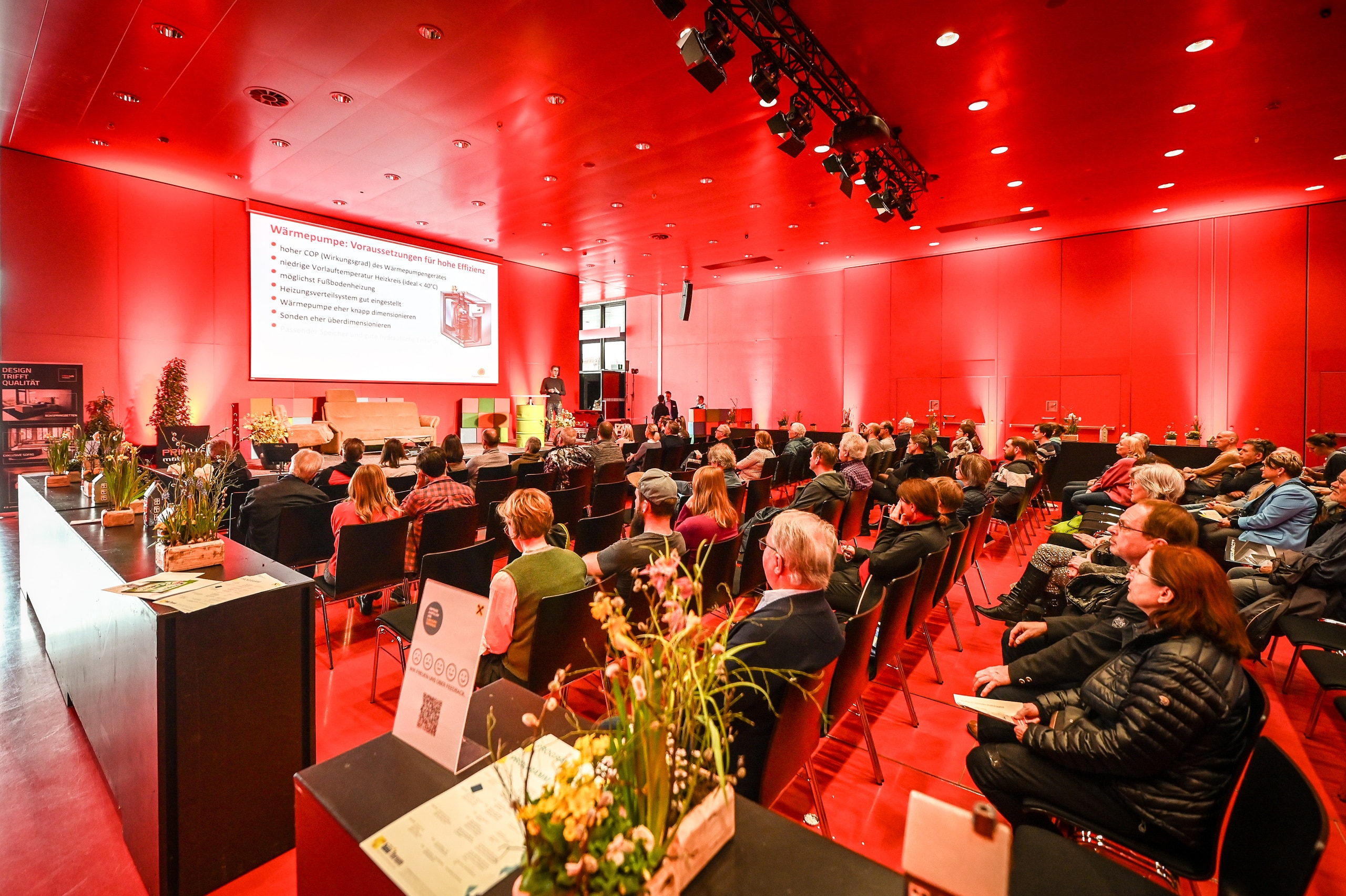 Ein roter Saal mit Podium, Präsentationsschirm und sitzender Zuhörerschaft auf der "com:bau" der Messe Dornbirn.