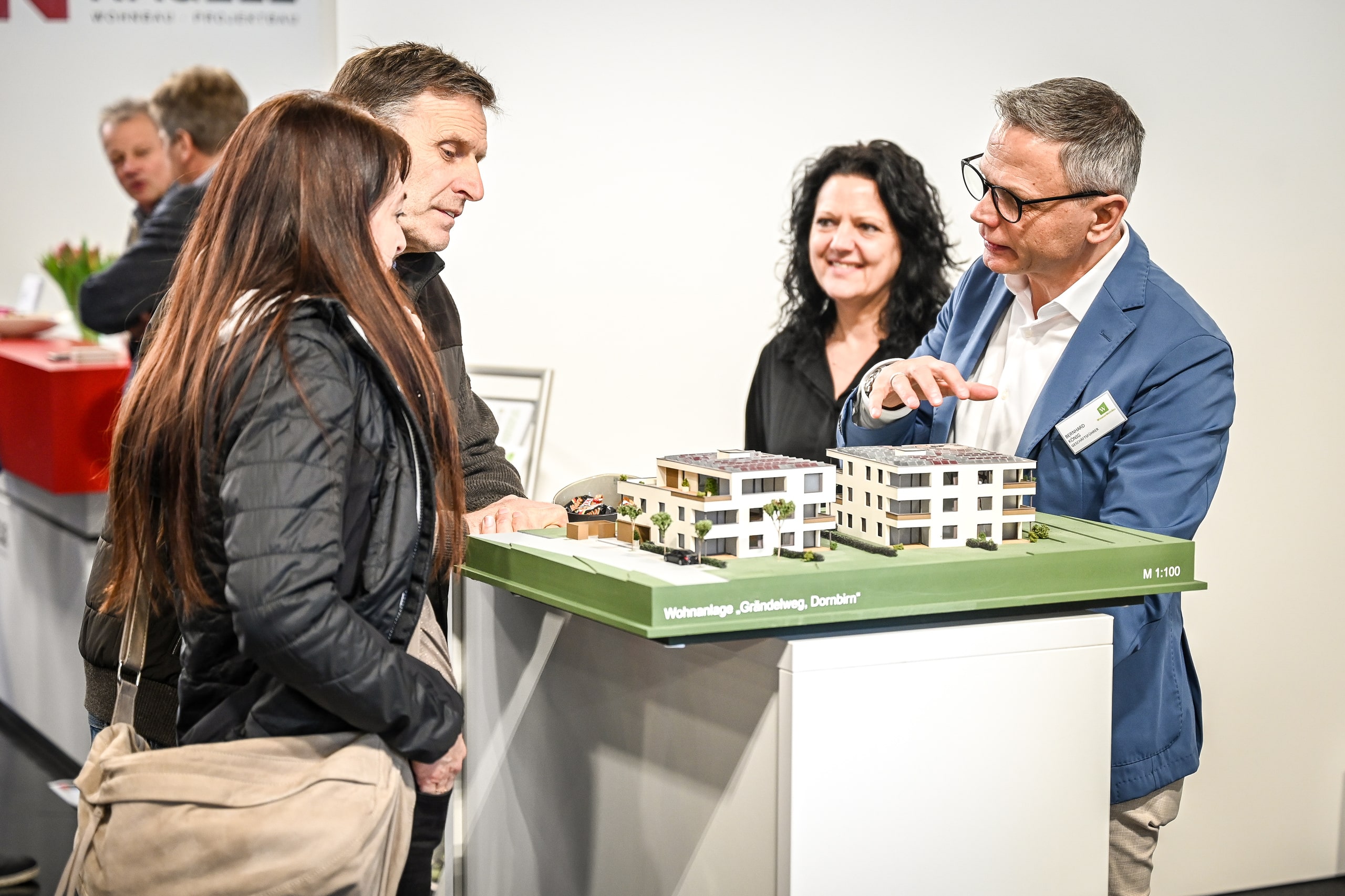 Ein Beratungsgespräch an einem Messestand der Messe com:bau mit einem Architektur-Hausmodell auf einem Tisch, bei dem Fachberater Besucher zu Planung und Kauf einer Wohnanlage oder eines Eigenheims informieren.