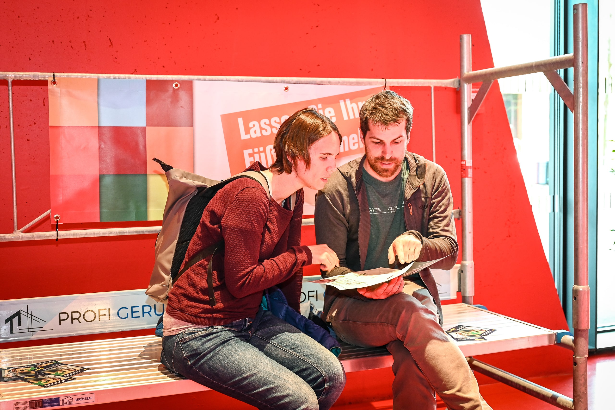Ein Paar mit Prospekt sitzend auf einem Baugerüst als Symbol für ein gemeinsames Bauprojekt, aufgenommen beim Rundgang durch die Ausstellung der Baumesse com:bau im Messequartier Dornbirn.