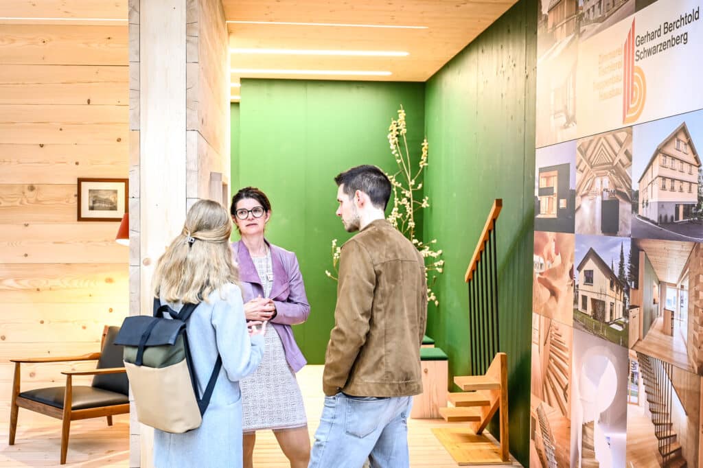 Ein Messestand mit Wohnraum‑Beratung, bei dem eine Wand mit dekorativer Holzvertäfelung im Fokus steht und ein Berater einer Besucherin Details zur Gestaltung und Materialwahl erklärt, aufgenommen auf der com:bau im Messequartier Dornbirn.
