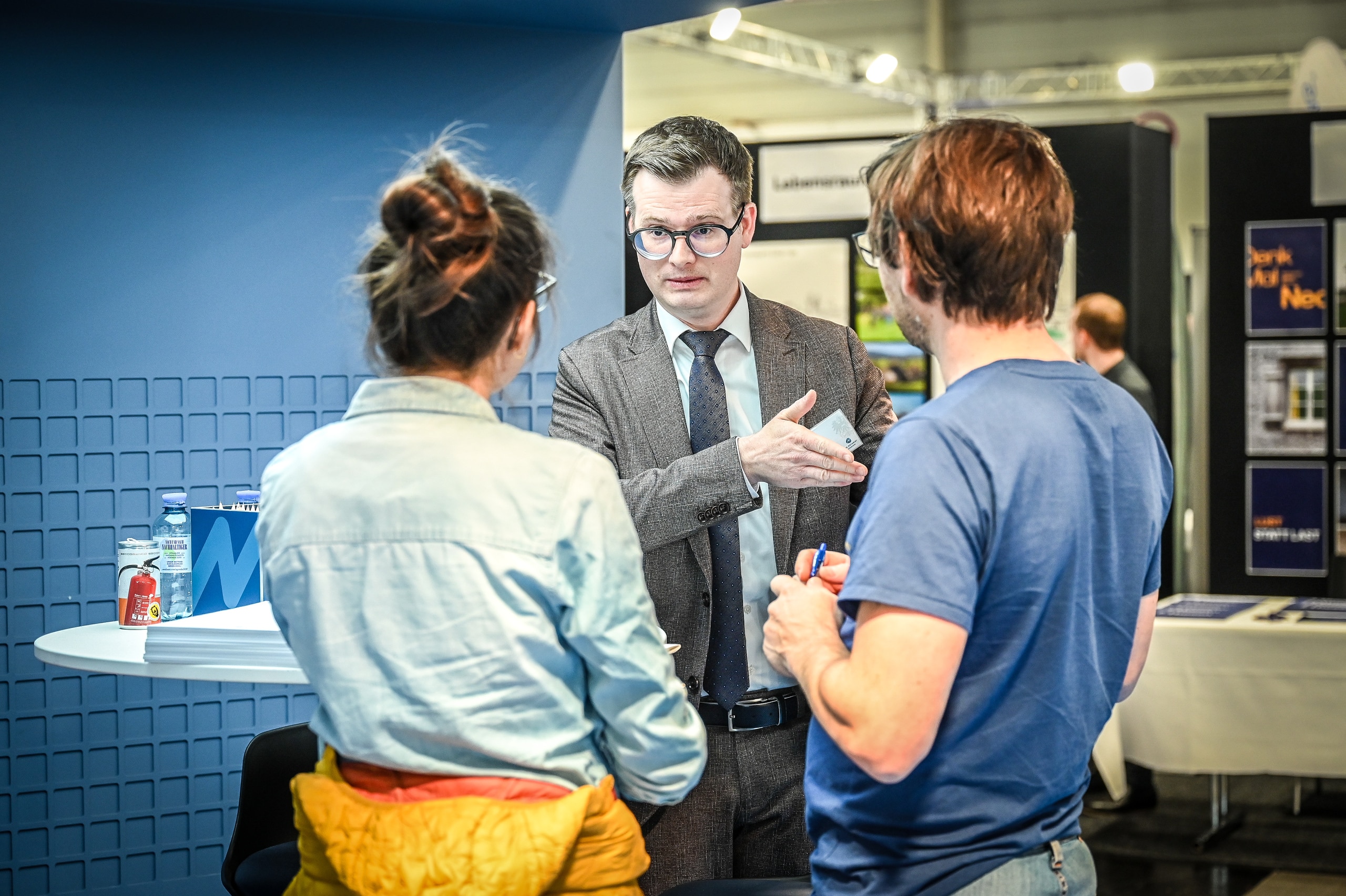 Ein Beratungsgespräch an einem Messestand mit einer modernen blauen Wandverkleidung mit einem Fachberater und einem Paar vor einem Tisch mit Prospekten und Materialmustern in einer Ausstellungshalle der Baumesse com:bau im Messequartier Dornbirn.