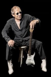 Adriano Celentano-Imitator Adolfo Sebastiani sitzt mit Sonnenbrille und einer weiss-schwarzen E-Gitarre vor schwarzem Hintergrund.