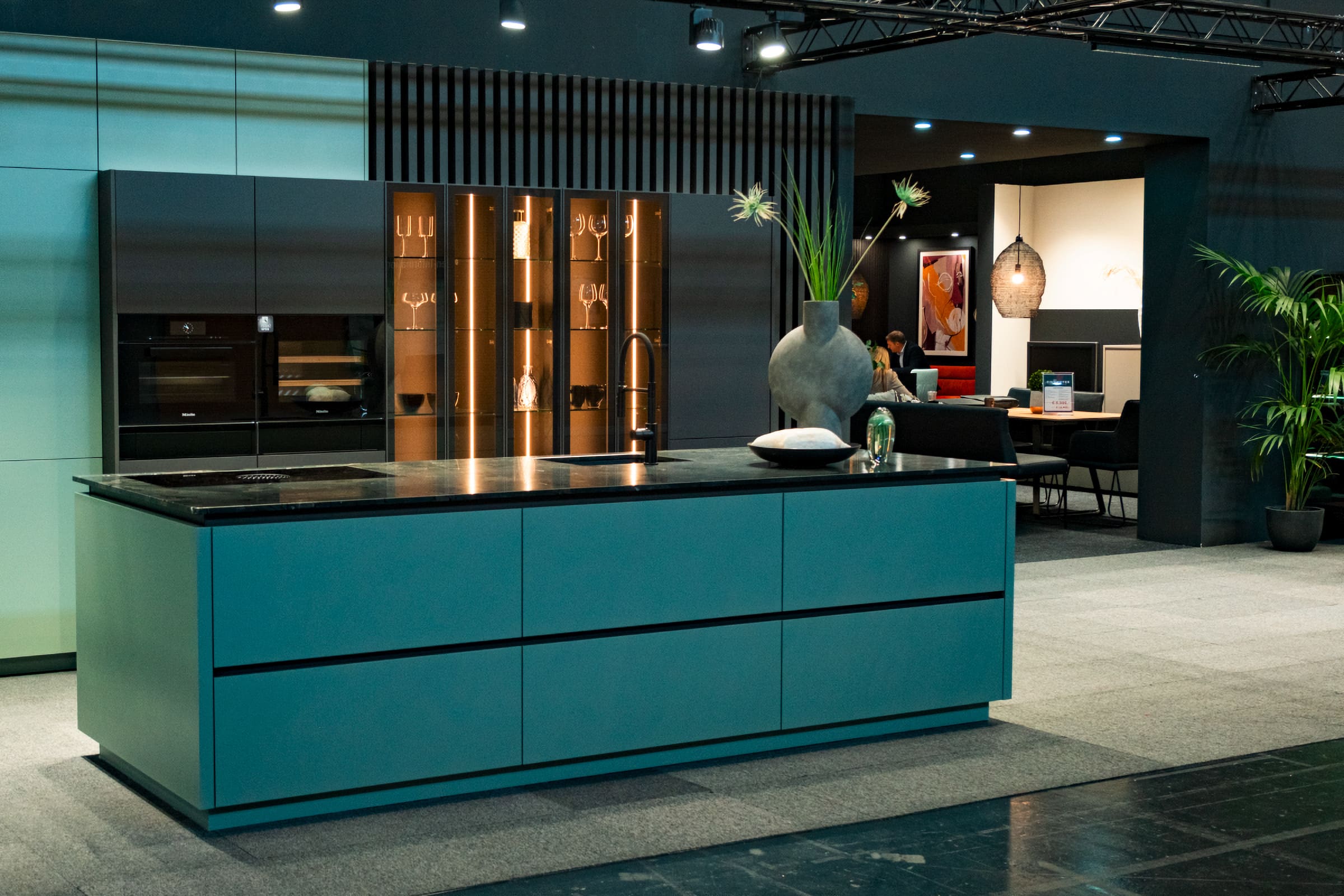 Eine Küche mit grifflosen Fronten, Kochinsel mit schwarzer Arbeitsfläche und Küchenzeile in schwarz mit integrierter beleuchteter Glasvitrine auf der Messe Wohnen & Interieur in der VIECON.