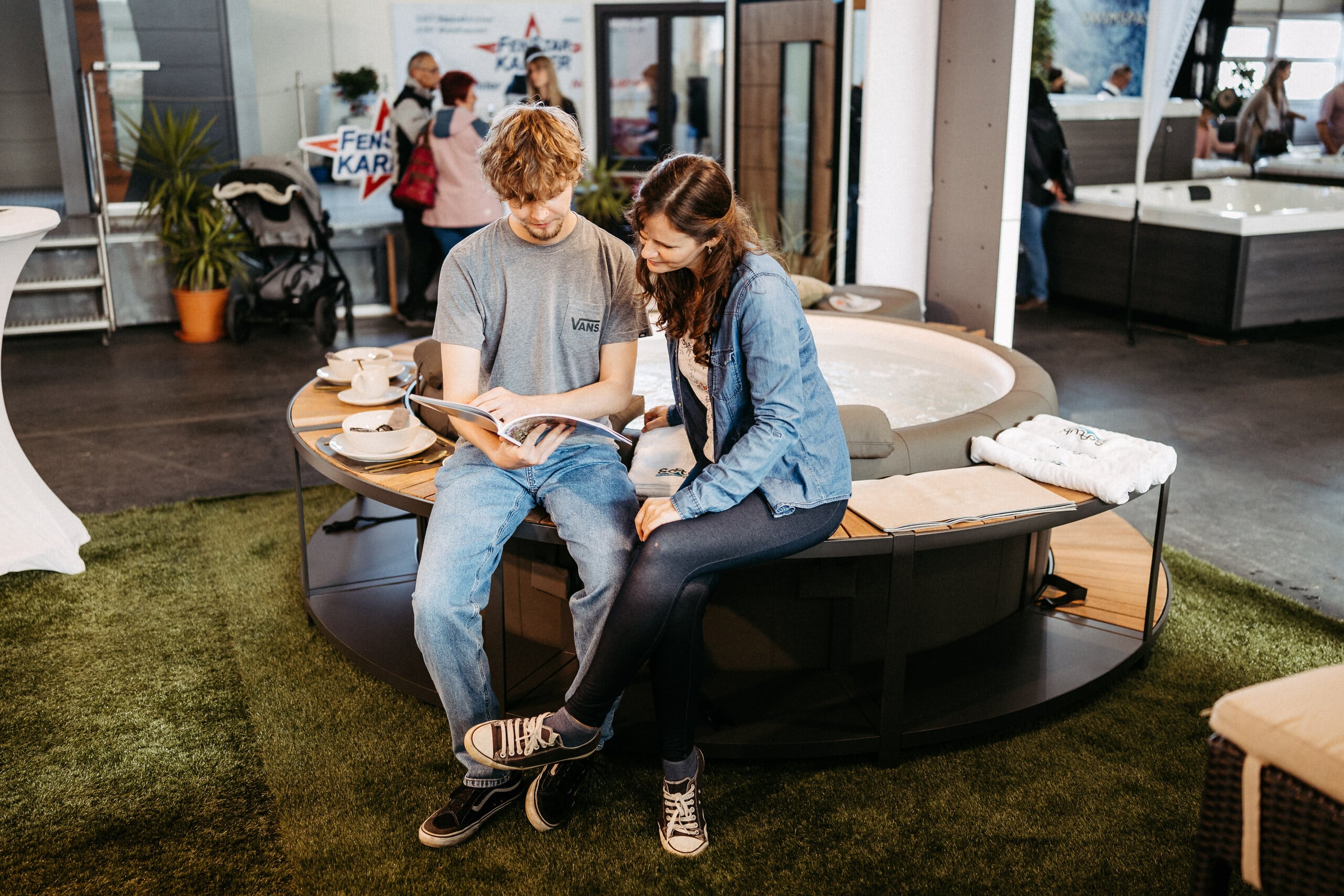 Ein Paar sitzt entspannt auf einer Bank vor einem Whirlpool und blättert gemeinsam in einem Produktkatalog in der Wellness-Sektion der Messe Wieselburg.