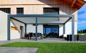 Eine moderne, anthrazitfarbene fassadenmontierte Lamellenpergola an mit gekippten weißen Lamellen und ohne Türelemente für einen luftigen Außenbereich der sich harmonisch in die Hausarchitektur einfügt von Moltke.