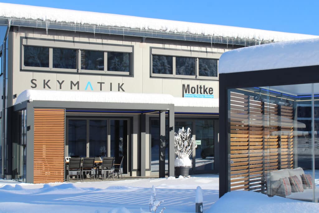 Eine moderne Terrasse im Winter mit einer freistehenden und einer hausmontierten Lamellenpergola mit Glasschiebetüren und Holzlamellenelementen von Moltke.