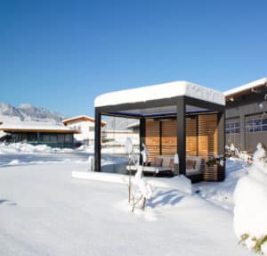 Eine moderne freistehende Lamellenpergola mit Holzrückwand, seitlich geschlossenen Glasschiebewänden, Outdoorcouch und Deckenbeleuchtung in winterlicher Schneelandschaft von Moltke.
