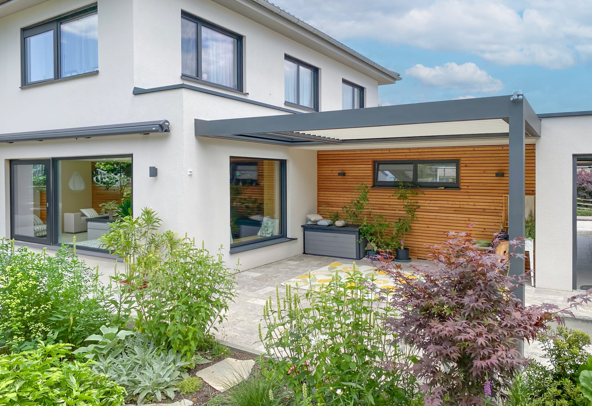 Eine moderne geflieste Terrasse mit Gartenfläche und einer hausmontierten Lamellenpergola mit weißen Lamellen und Gestell in Anthrazit sowie eingezogener Markise in gleicher Farbe von Moltke.