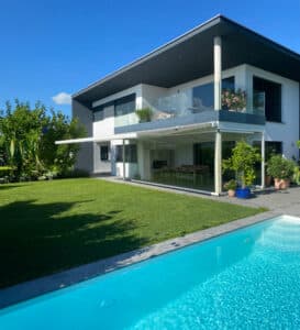Ein modernes Einfamilienhaus mit Balkon, Wintergarten mit Glasschiebetüren, großem Garten mit Pool und einer fassadenmontierten weißen Gelenkarmmarkise von Moltke.