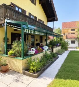 Bäuerliches Haus im Altbau mit klassischer Terrasse und dunkelgrüner Pergola mit Schrägdach und gelb-grün gestreiften Markisen in sonniger Gartenatmosphäre.