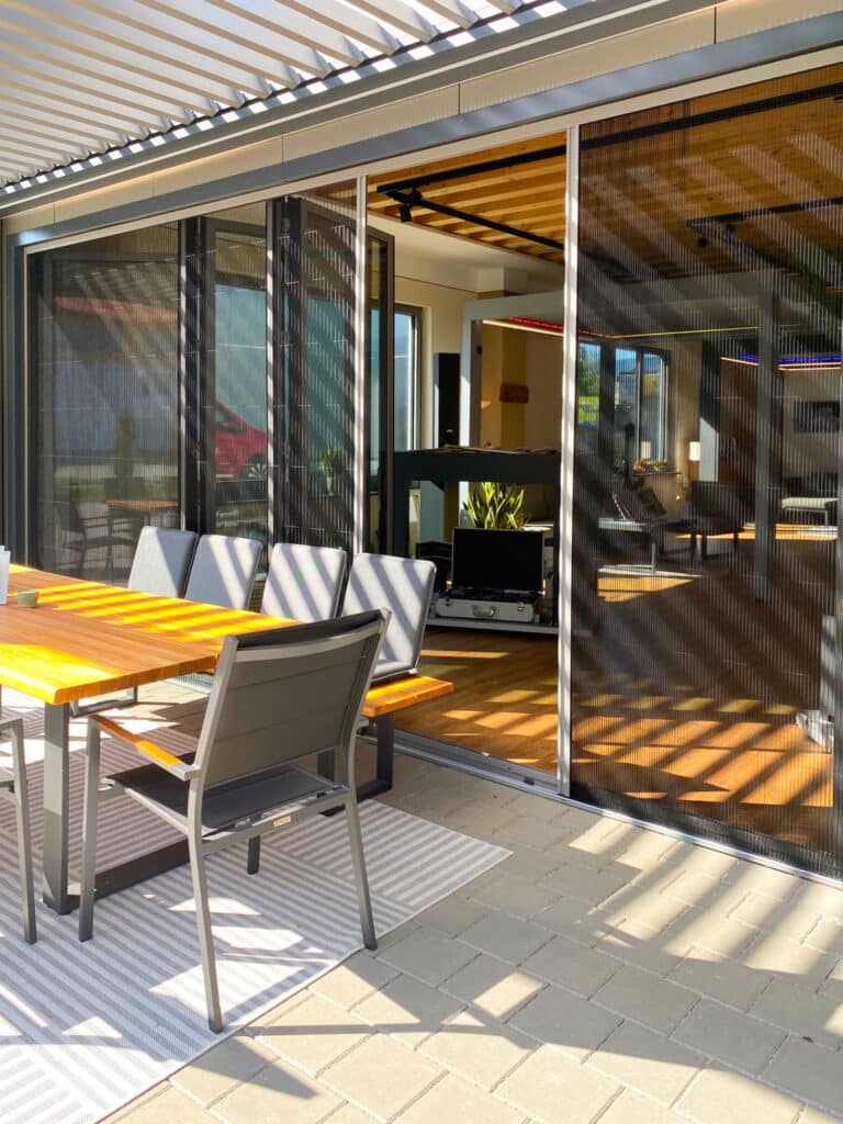 Terrasse mit halboffenen Insektenschutz-Schiebetüren unter einer modernen Lamellenpergola und Glasfaltwänden als Terrassentüren von Moltke.
