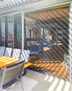 Seitenaufnahme einer Terrasse unter einer modernen Lamellenpergola mit geschlossenen Insektenschutz-Schiebetüren und Glasfaltwänden als Terrassentüren von Moltke.