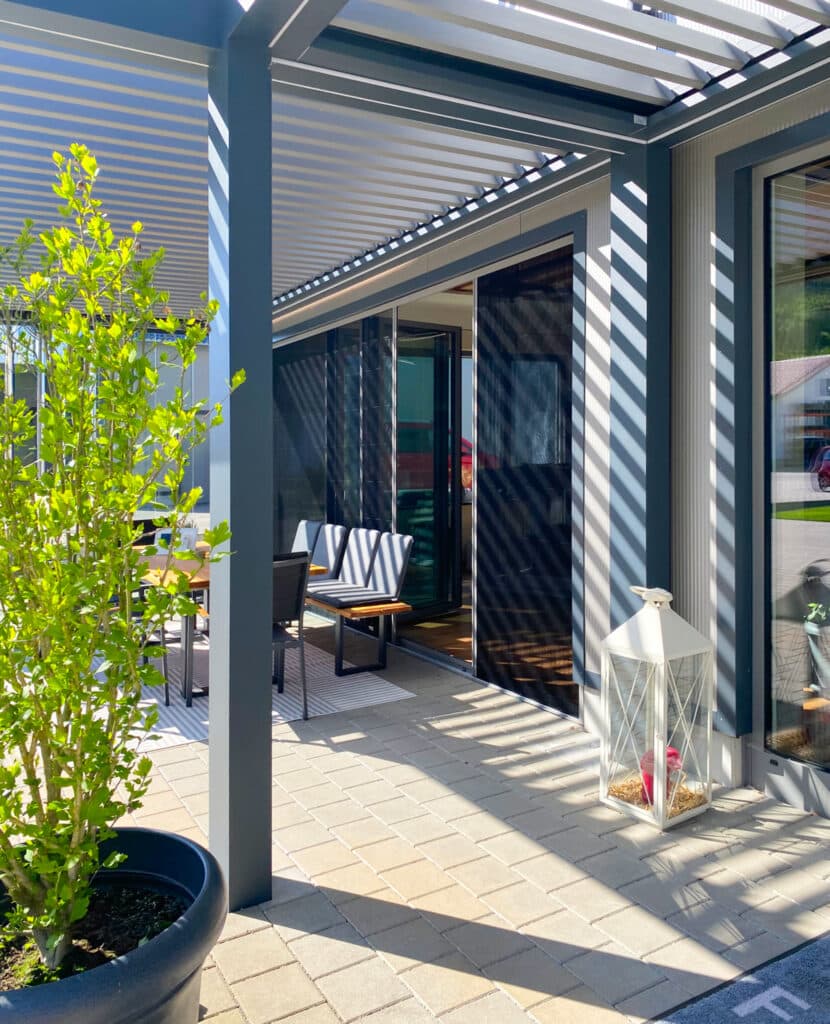 Seitenaufnahme einer Terrasse unter einer modernen Lamellenpergola mit halboffenen Insektenschutz-Schiebetüren und Glasfaltwänden als Terrassentüren von Moltke.