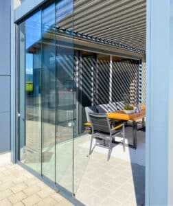 Seitenaufnahme einer Terrasse mit halboffenen Glasschiebe-Seitenelementen unter einer modernen Lamellenpergola mit geschlossenen Insektenschutz-Schiebetüren und Glasfaltwänden als Terrassentüren von Moltke.
