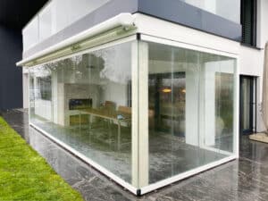 Eine moderne Pergola bzw. Wintergarten mit weißem Rahmen und eleganten geschlossenen Glasschiebe-Elementen, einem darüberliegendem Balkon und einer eingezogenen Markise von Moltke.
