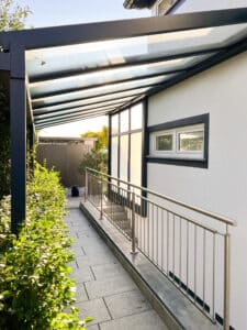 Ein modernes Haus mit einem eleganten, schräg verlaufenden Glasvordach im Pergola-Stil in anthrazitfarbener Rahmenkonstruktion von Moltke, das als stilvoller und funktionaler Wetterschutz für den Eingangsbereich dient und sich harmonisch in die Architektur einfügt.