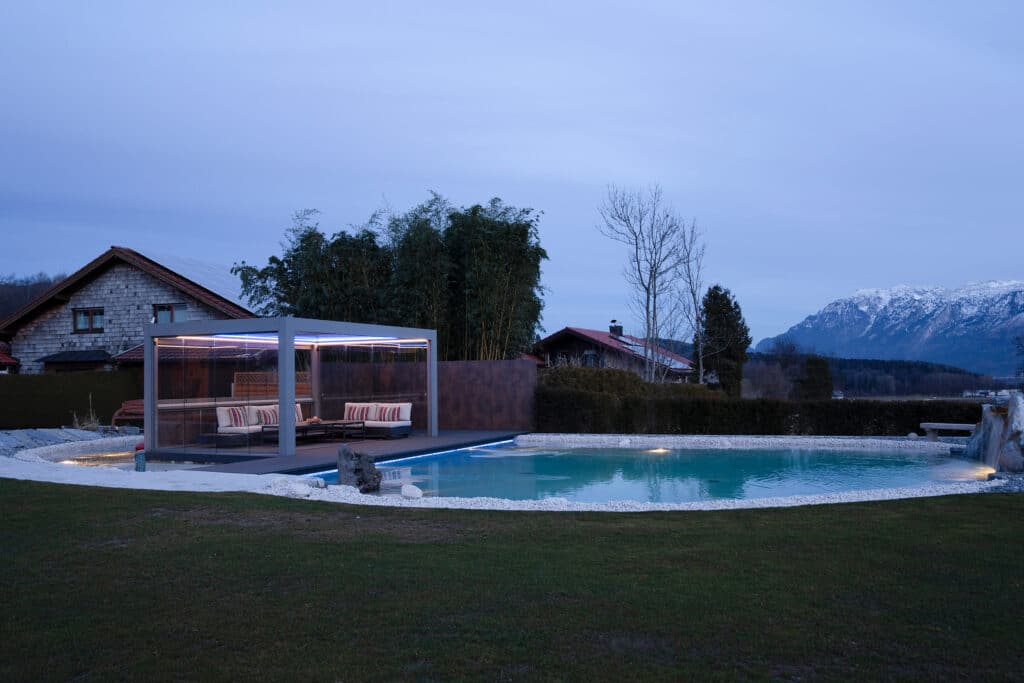 Ein moderner Garten in Abendstimmung mit Schwimmteich und hellgrauer Pergola mit geschlossenem beleuchtetem Lamellendach und geschlossenen Glasschiebetüren von Moltke.