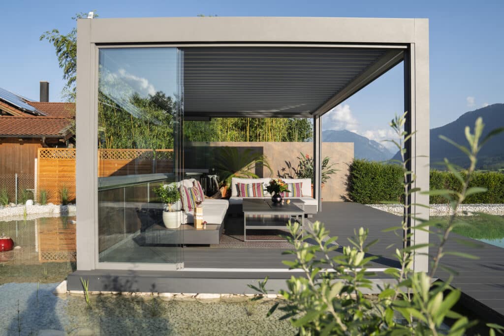 Ein moderner Garten mit Teich und einer hellgrauen Pergola mit Lamellendach in seitlicher Ansicht mit offenen Glasschiebetüren von Moltke.