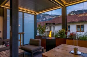 Ein Wintergartenanbau in Anthrazit mit geschlossenem, indirekt mit LED beleuchtetem Lamellendach und teilweise geschlossenen Glasschiebetüren in gemütlicher Abendstimmung mit brennender Feuersäule von Moltke.
