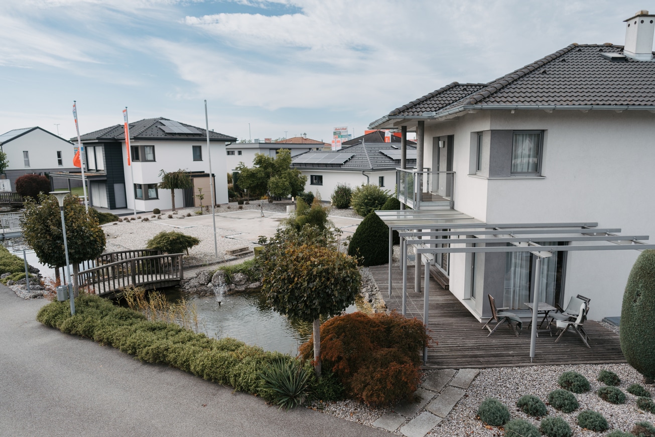 Blick auf ein Einfamilienhaus mit Holzterrasse und überdachter Pergola mit Teich im Musterhauspark Haid.