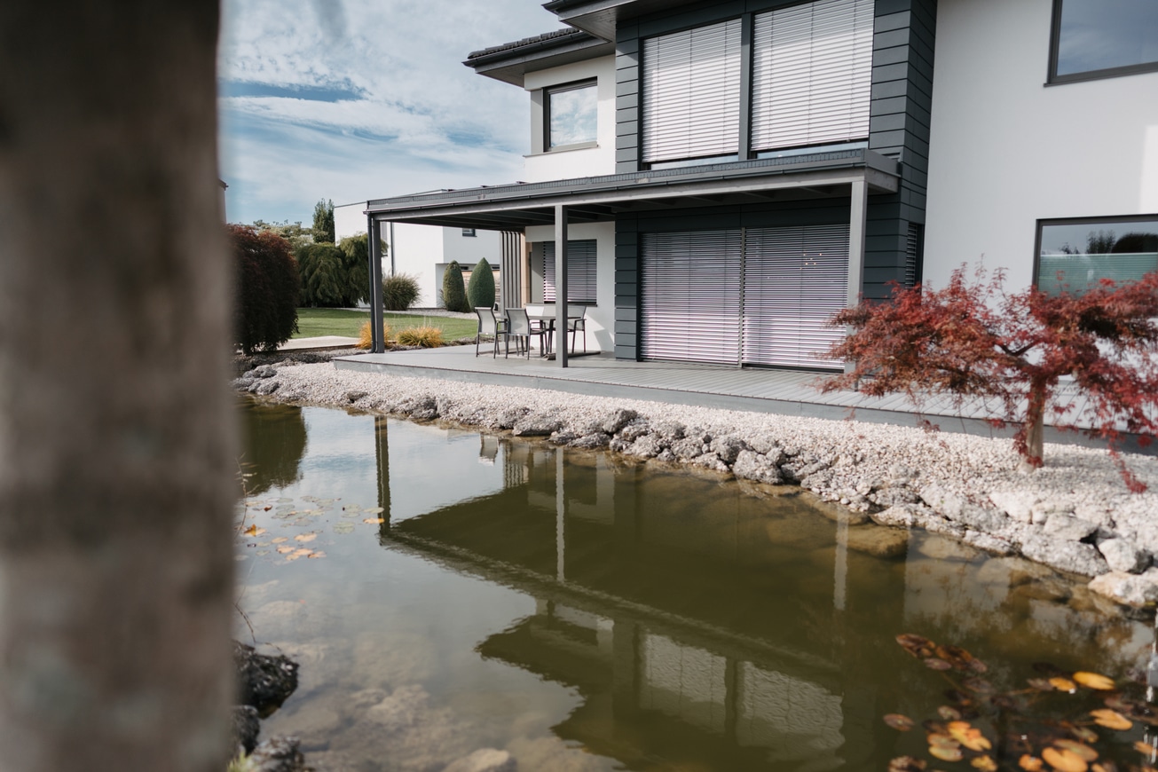 Einfamilienhaus mit überdachtem Terrassenbereich, Fassadenverkleidungselementen in Schwarz und vorm Haus liegendem Teich im Musterhauspark Haid.