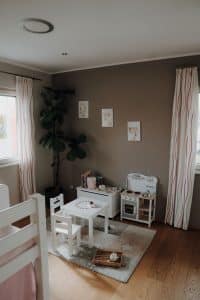 Ein geräumiges Kinderzimmer mit Bett, zwei Fenstern, einer Spielküche, Kommode, einer XXL-Pflanze und einem Maltisch mit zwei Stühlen im Musterhauspark .