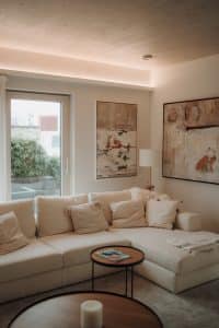 Ein einladendes helles Wohnzimmer mit weisser Couch, freiliegendem grauem Hochflorteppich, Couchtischensemble, Terrassentür, zwei Gemälde in Naturtönen, einer Stehleuchte, Wandleiste mit indirektem warmen Raumlicht und einer Holzdecke im Musterhauspark.