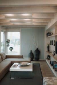 Offener Wohnraum mit Eckfenster und Glastür zur Terrasse, einem L-förmigen Sofa in Grau, Couchtisch, beigem Fliesenboden, freiliegendem Teppich, einer eingekofferten, gemauerten Medienwand und einer hellen Holzbalkendecke mit indirekter Beleuchtung im Musterhauspark.