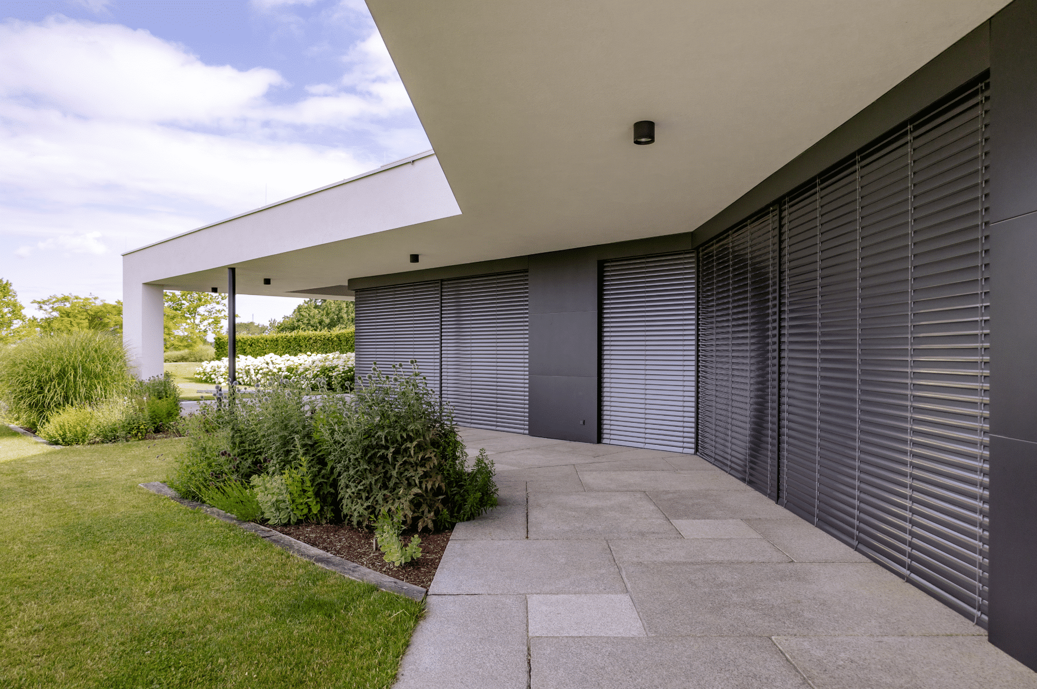 Newo Sonnen- und Insektenschutz zeigt ein modernes Einfamilienhaus mit großen Glasfronten auf denen Rollläden als Sonnenschutz angebracht wurden und einer überdachten Terrasse.
