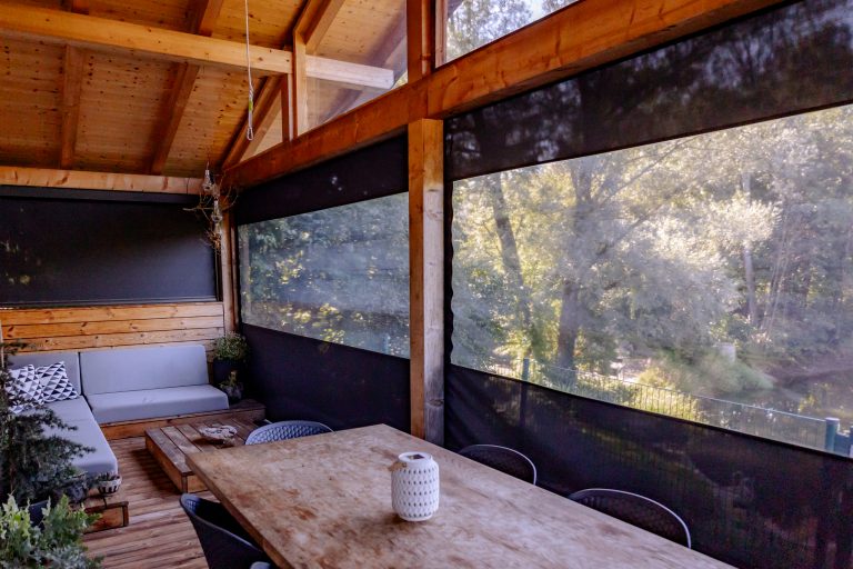 Newo Sonnen- und Insektenschutz zeigt eine überdachte Terrasse eine Holzhauses, die mit durchsichtigen Textilscreens geschlossen werden kann.