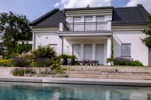 Newo Sonnen- und Insektenschutz zeigt ein Einfamilienhaus mit Terrasse und Balkon und bodentiefen Fenstern, die mit weißen Raffstores beschattet werden.