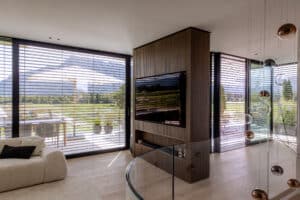 Ein Wohnzimmer mit Eckverglasung und Terrassenschiebetüren mit schwarzem Alu-Rahmen und halboffenen Raffstores als Sonnen- und Hitzeschutz von Newo.