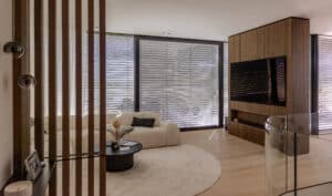 Ein Wohnzimmer mit Eckverglasung und Terrassenschiebetüren mit schwarzem Alu-Rahmen welches trotz der geschlossenen silbernen Raffstores hell und freundlich ist, von Newo.