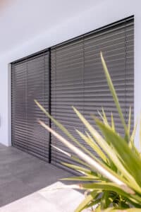 Eine Terrassenschiebetür bzw. Wandverglasung mit schwarzem Alu-Rahmen und außenliegenden, dunkelgrauen, geschlossenen Raffstores von Newo.