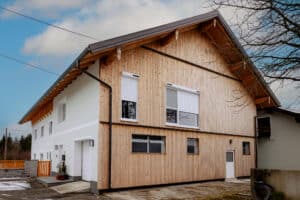 Ein Haus mit Satteldach, einseitiger Holzfassadenverkleidung und Fenstern mit Vorbaurollladen in Weiss mit Solarmotor von NEWO.