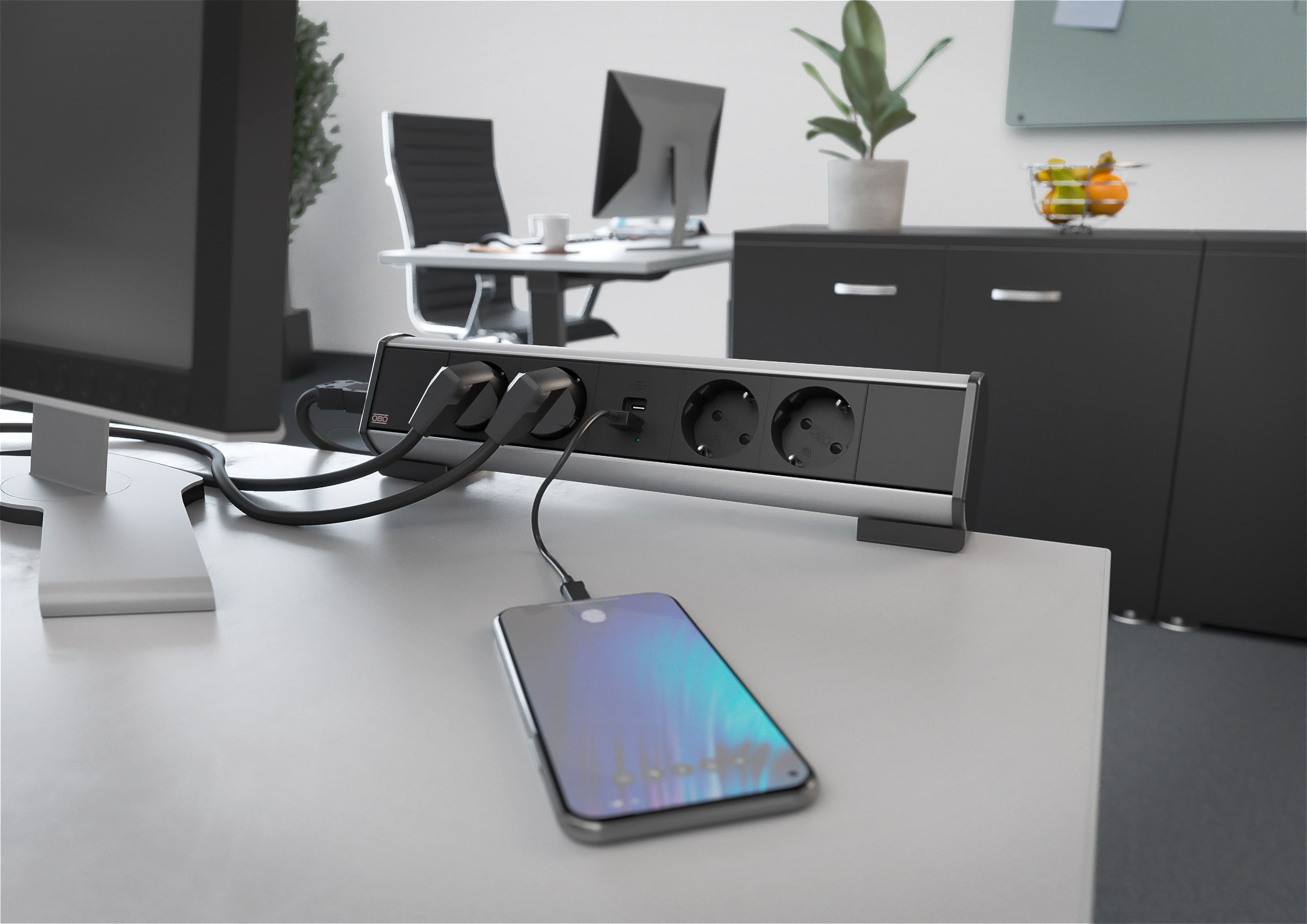 OBO Bettermann zeigt einen Arbeitsplatz mit schwarz-grauer Steckdosenleiste mit USB-Charger und Befestigungszwinge am Schreibtisch.