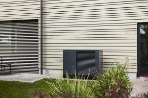 Schwarz-dunkelgraue Wärmepumpe von ÖkoFEN in einem Garten vor einem Einfamilienhaus mit heller Holzfassade in Sprossenoptik.