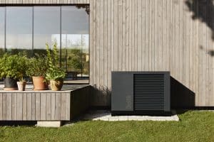 Schwarz-dunkelgraue Wärmepumpe von ÖkoFEN vor einem Haus mit Holzfassade in Sprossenoptik und einem erhöhten Terrassenpodest mit Blumentöpfen.