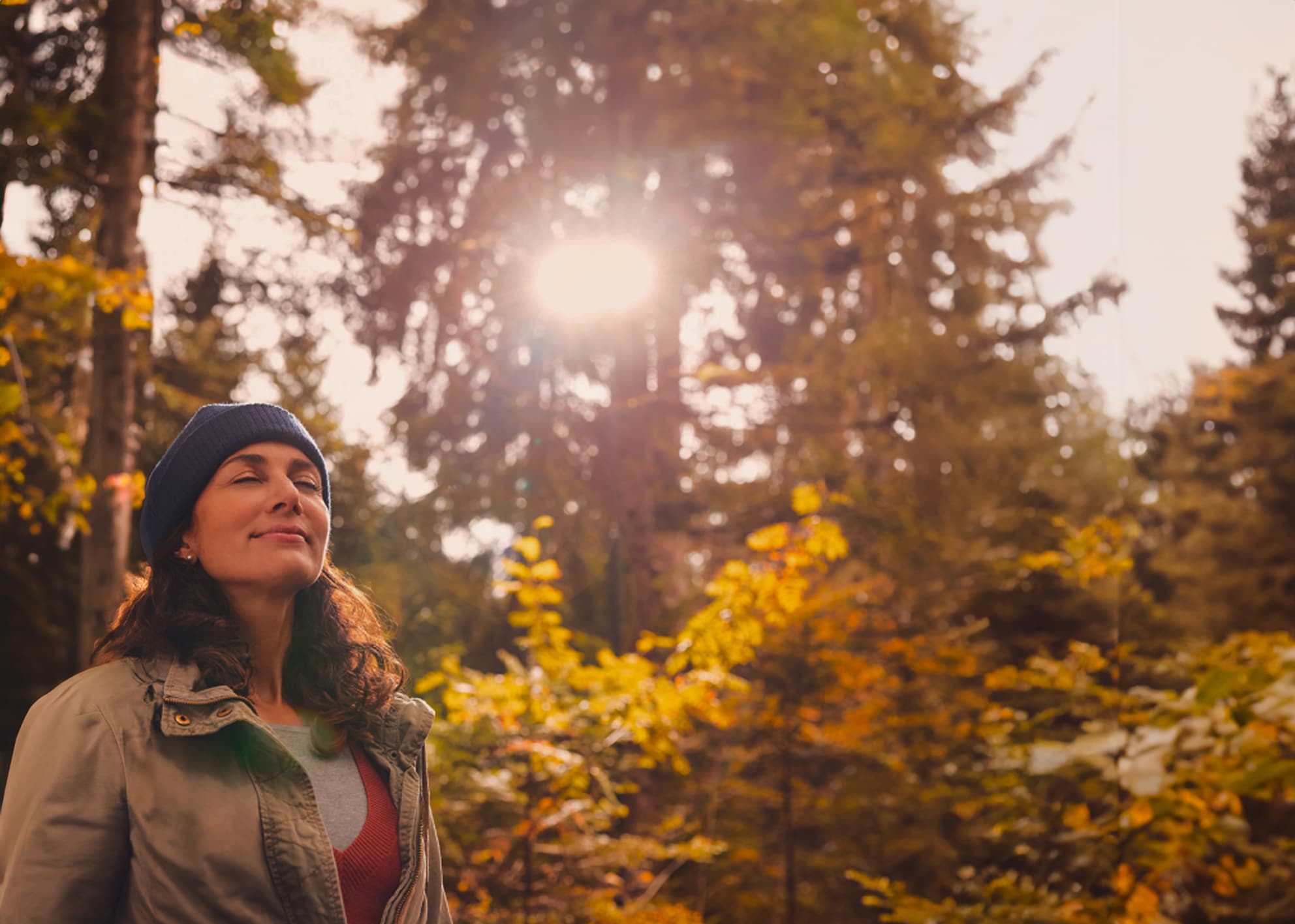 Eine Frau steht im Wald und genießt die Herbstsonne die durch die Bäume strahlt.