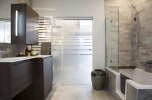 Modernes barrierefreies Badezimmer mit Möbeln aus dunklem Holz, Boden- und Wandfliesen in Steinoptik, Glas-Schiebetür und einer Badewanne mit Seiteneinstieg von HOLTER gezeigt von Limbach.