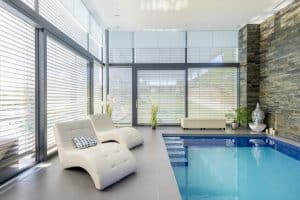 Private Schwimmhalle mit Indoorpool und geschwungenen Relaxliegen, Wandfliesen in Steinoptik und zum Garten führenden Hebeschiebetüren mit Jalousien von Planungsbüro Schaufler.
