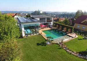 Villa mit Flachdach, dachmontierten Solarpaneelen, privatem Hallenbad, einem großzügigem Garten, Terrasse mit Fliesen in Holzoptik und Naturpool aus der Vogelperspektive von Planungsbüro Schaufler.